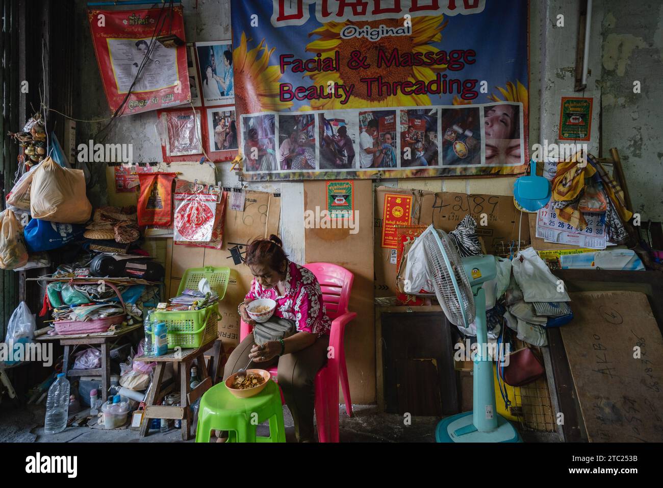 Bangkok, Thailandia. 8 dicembre 2023. Una nonna thailandese è vista pranzare davanti alla pubblicità per un salone di bellezza di strada per il threading di bellezza del viso e massaggi, situato all'entrata di un santuario in Charoen Krung Road. Chiamato "mang ming" in tailandese, il threading del viso è un antico metodo di depilazione originario della Cina, dell'India e del mondo arabo. Il metodo utilizza fili di cotone sottili come alternativa alla ceretta o alla pinzatura delle sopracciglia sottili e rimuove i peli superflui del viso. Credito: SOPA Images Limited/Alamy Live News Foto Stock