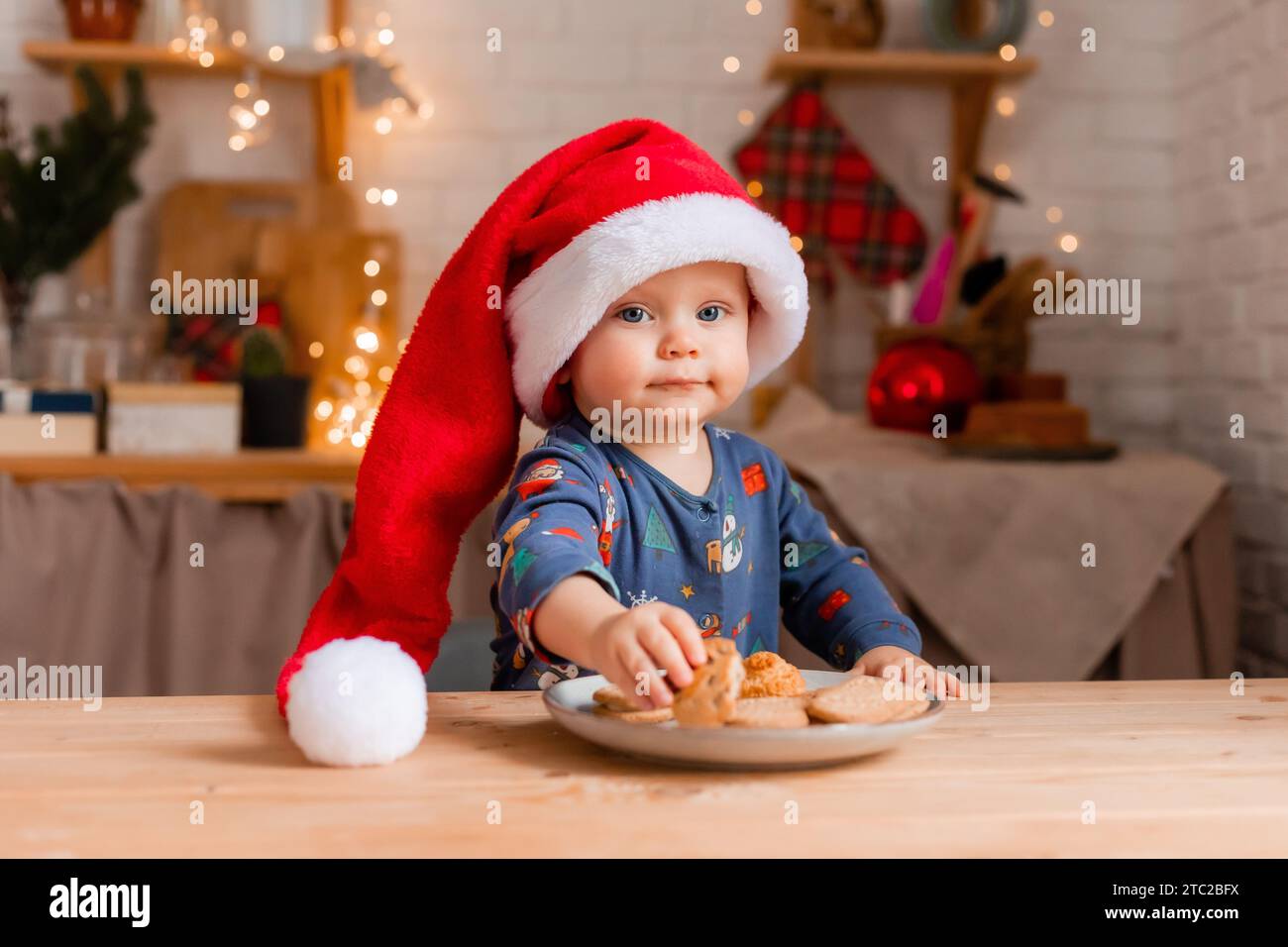 Un bambino carino in un cappello di Babbo Natale in cucina che mangia biscotti il giorno di Natale. Foto di alta qualità Foto Stock