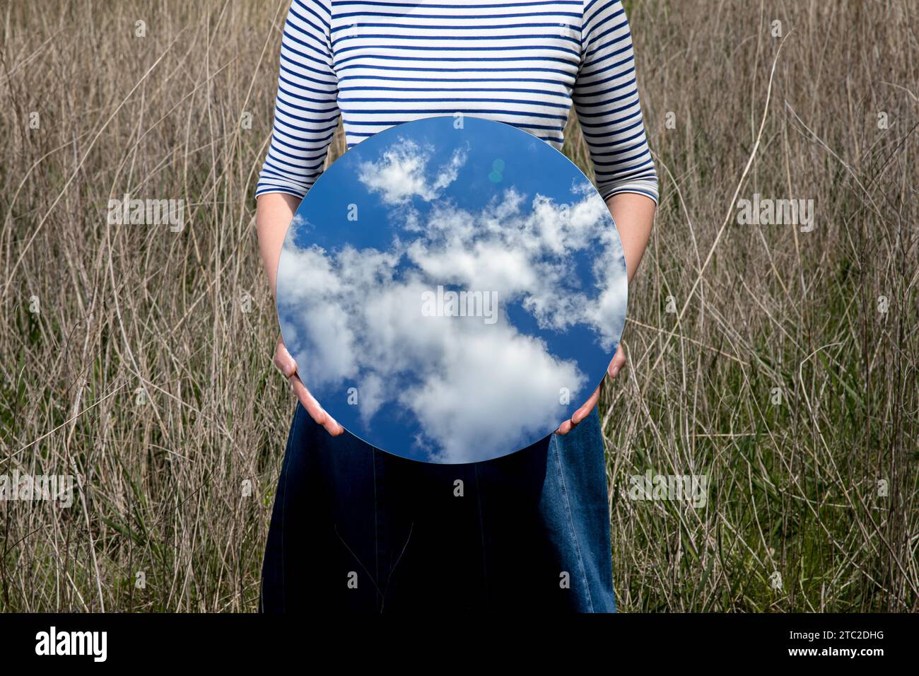 Surrealismo con una donna che tiene uno specchio e copre il suo volto nel campo con uno sfondo trasparente dietro lo specchio Foto Stock