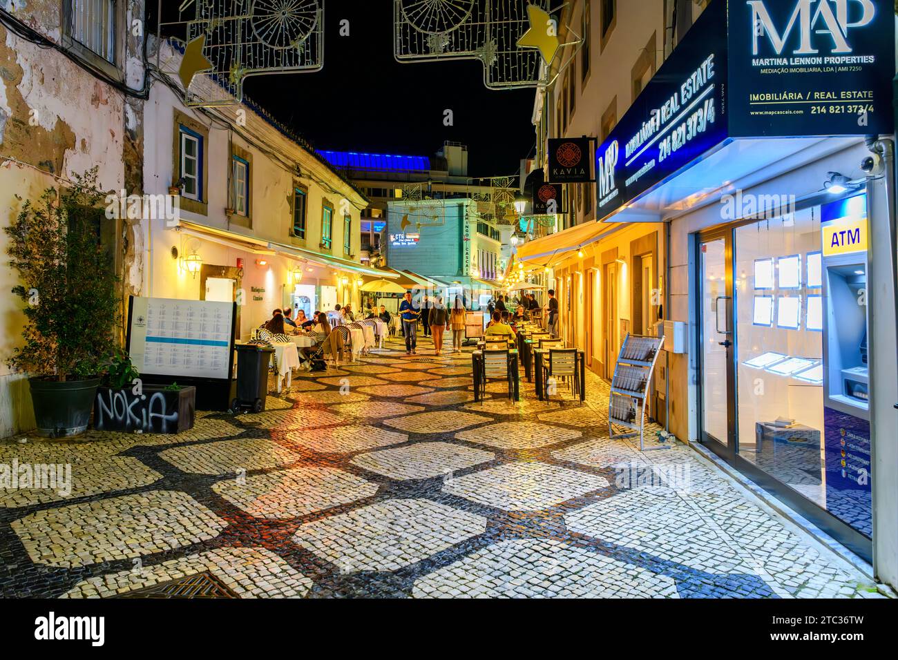 Una strada colorata di negozi e caffetterie con la pavimentazione di Calcada illuminata di notte, nella città vecchia della località balneare di Cascais in Portogallo. Foto Stock