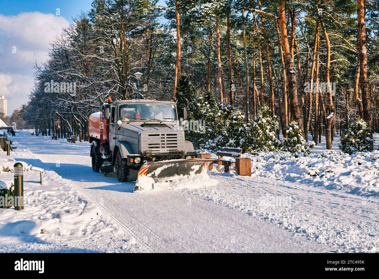 Una soleggiata giornata invernale nel parco cittadino, dove un vomere innevato ripulisce efficacemente la neve dai marciapiedi. Gli alberi innevati creano una pittoresca sce Foto Stock