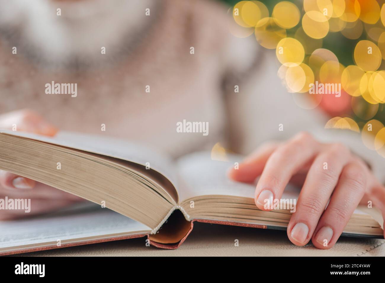 Libri di Natale.le mani in un maglione girano le pagine di un libro su un albero di Natale con sfondo ghirlande.lettura accogliente nella stagione invernale. Foto Stock