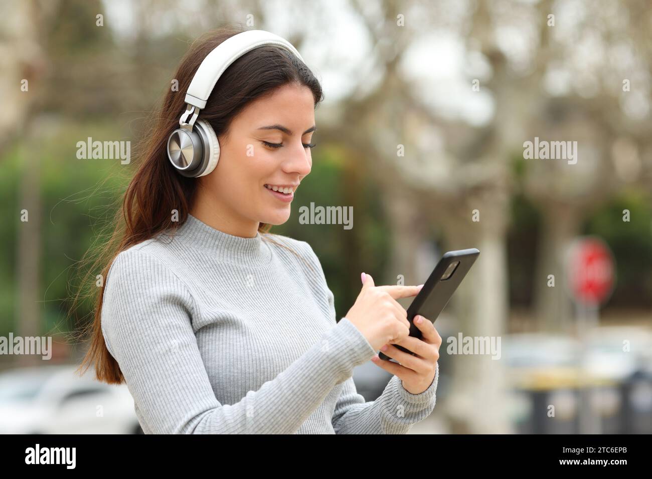 Donna che indossa cuffie che cammina per strada e controlla la musica al telefono Foto Stock