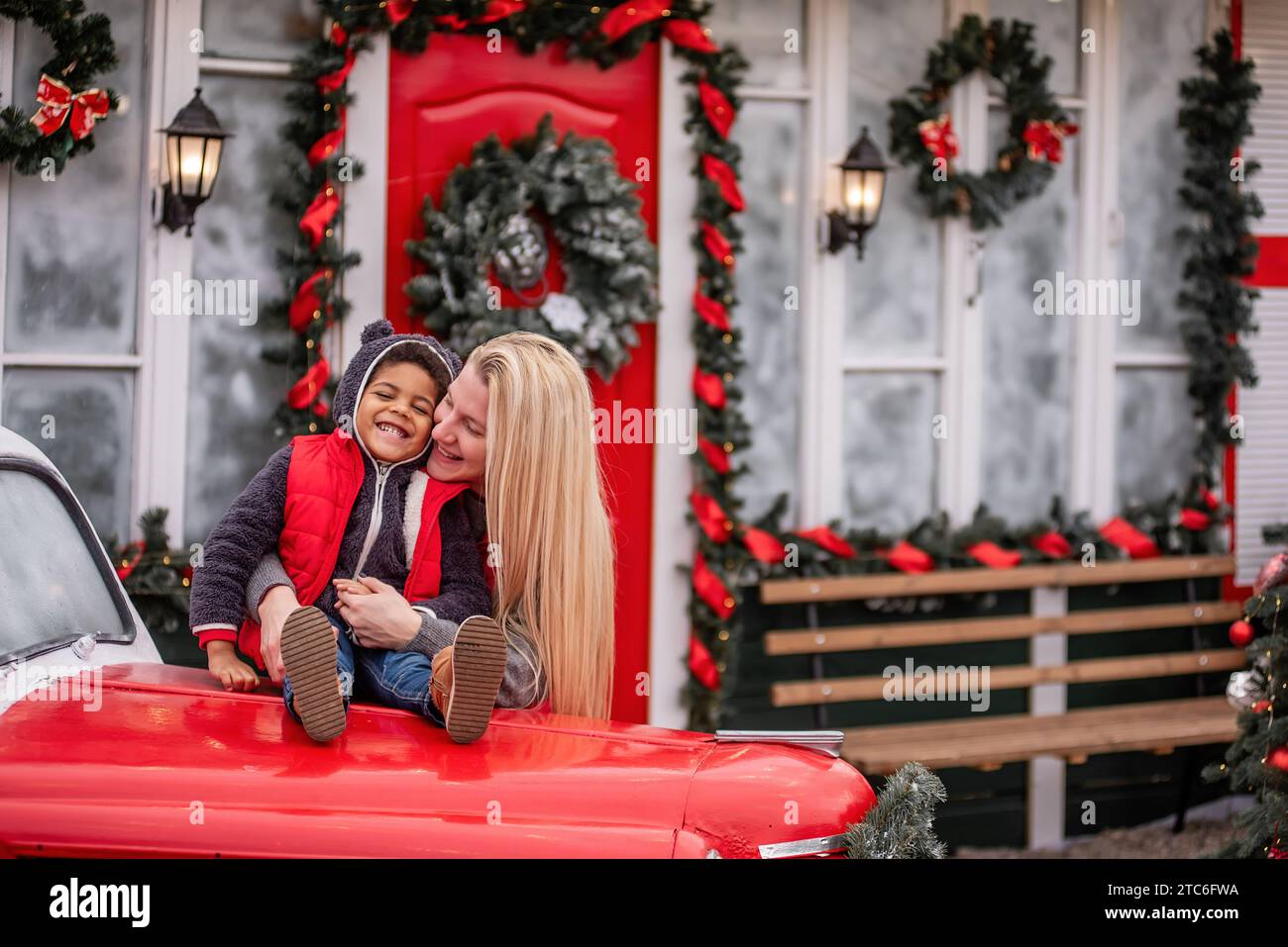 Un bambino afro-americano con madre caucasica siede sul cofano di un'auto d'epoca natalizia. Bambino con donna bionda che abbraccia, ingannando vicino al Foto Stock