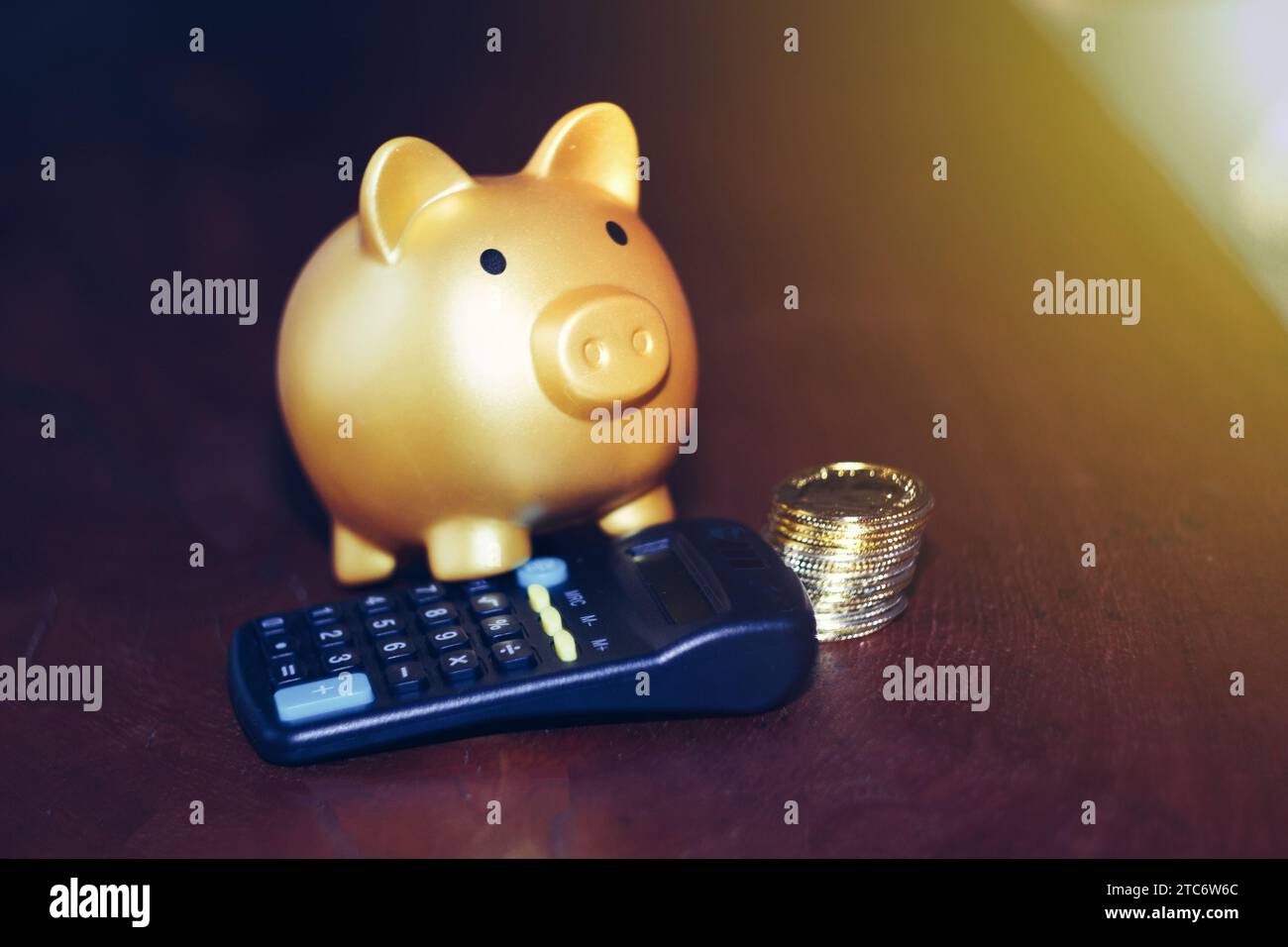 Un salvadanaio d'oro su una calcolatrice nera con monete sparse su un tavolo di legno scuro. Risparmio di finanziamenti, denaro ed economia. Foto Stock