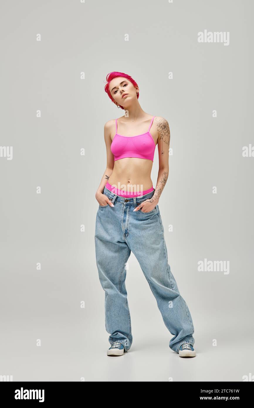 donna seducente con capelli rosa corti e tatuaggi in top rosa e jeans con le mani nelle tasche Foto Stock