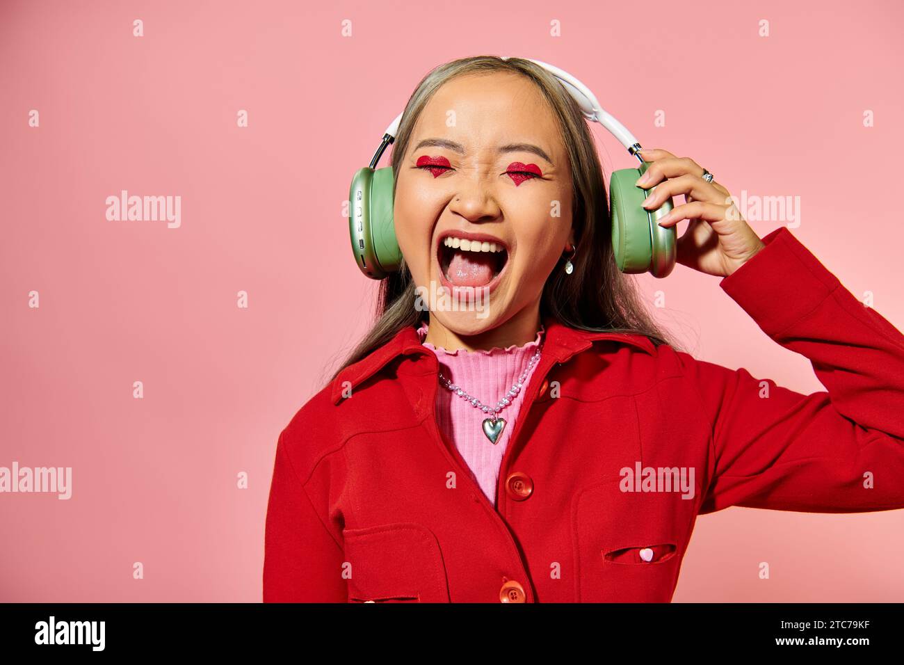 eccitata giovane donna asiatica che ascolta musica con cuffie wireless e urla su sfondo rosa Foto Stock