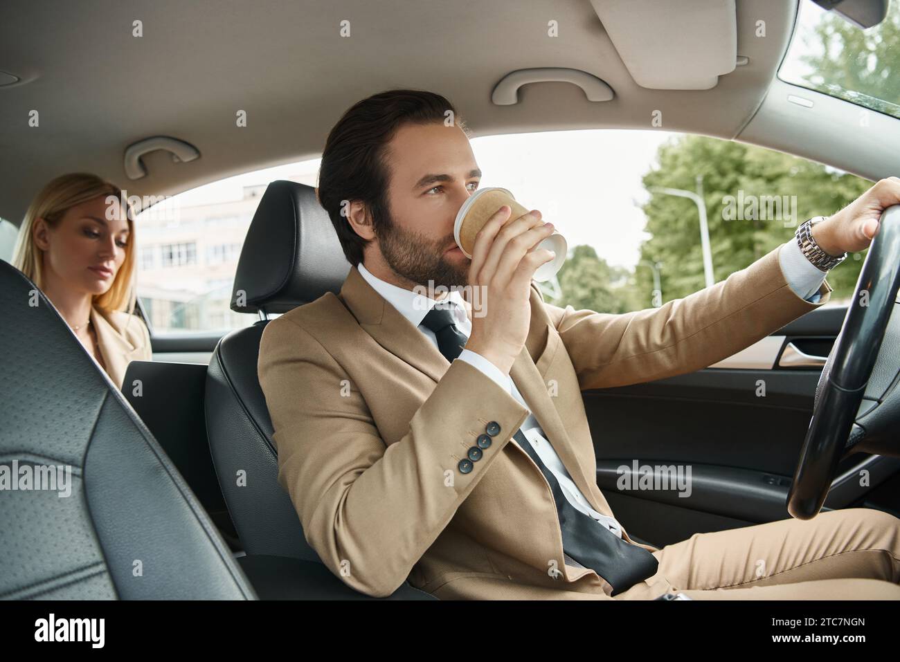 un uomo elegante e formale indossa un caffè e guida un'auto con una donna d'affari bionda sul sedile posteriore Foto Stock
