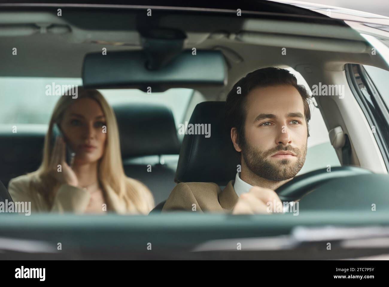 uomo barbuto concentrato che guida un'auto di lusso con una donna d'affari bionda che parla al telefono cellulare Foto Stock
