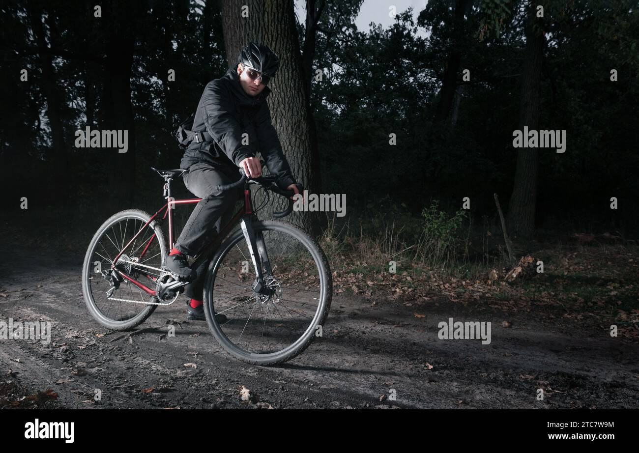 Ciclista in casco, su una bicicletta di ghiaia in piedi su un sentiero attraverso la foresta di sera. Foto Stock