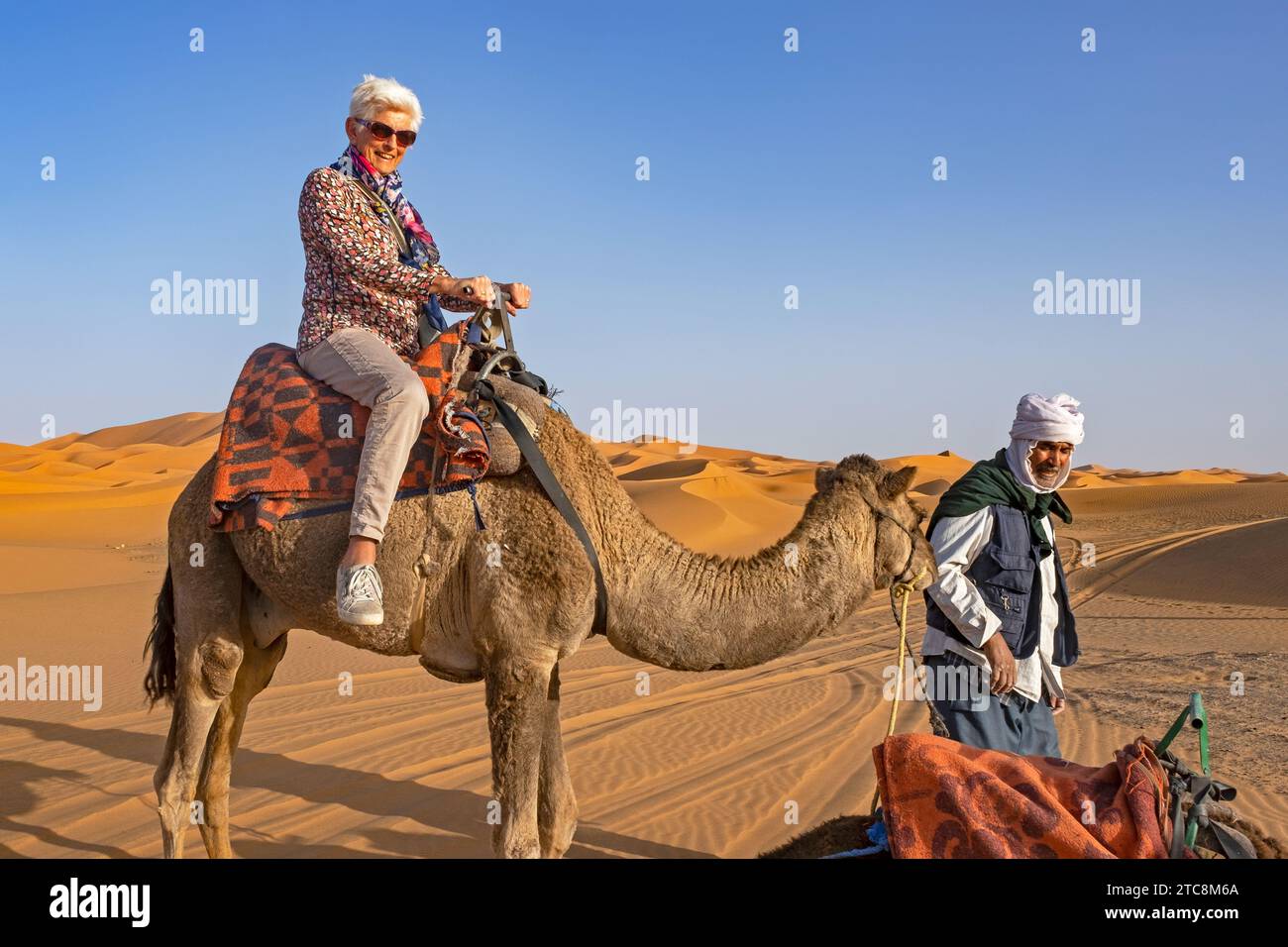 Cammellatore e turista occidentale cavalcando un cammello in una duna di sabbia di Erg Chebbi, deserto del Sahara vicino a Merzouga, Drâa-Tafilalet, Errachidia, Marocco Foto Stock