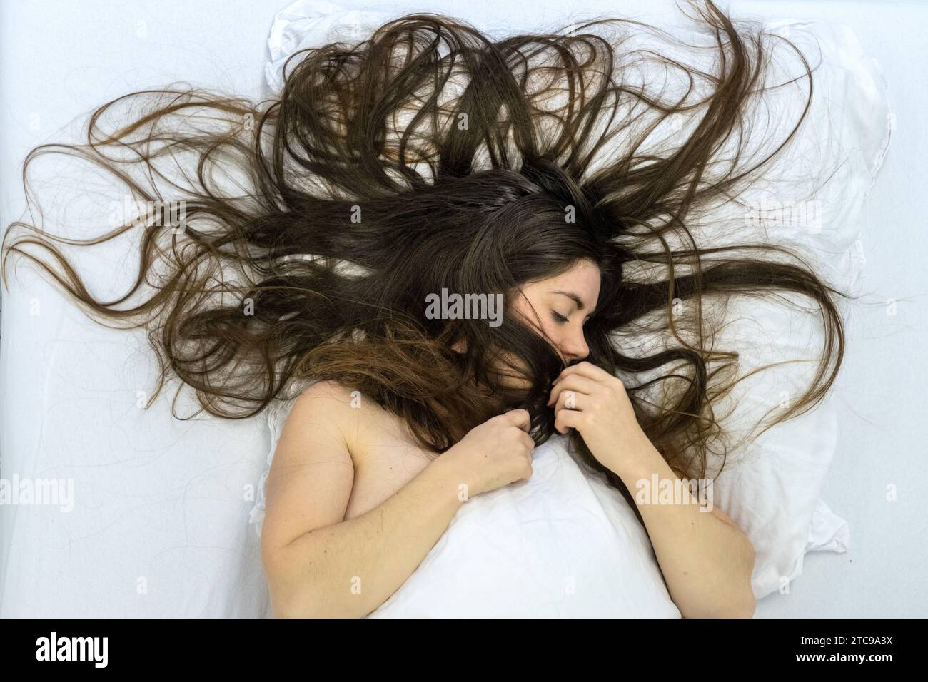 vista dall'alto di una seducente donna bruna nuda attraente, giovane, sexy e sensuale, lunghi capelli castani ricci selvaggi sulle lenzuola, la domenica mattina, sdraiata a letto, Foto Stock