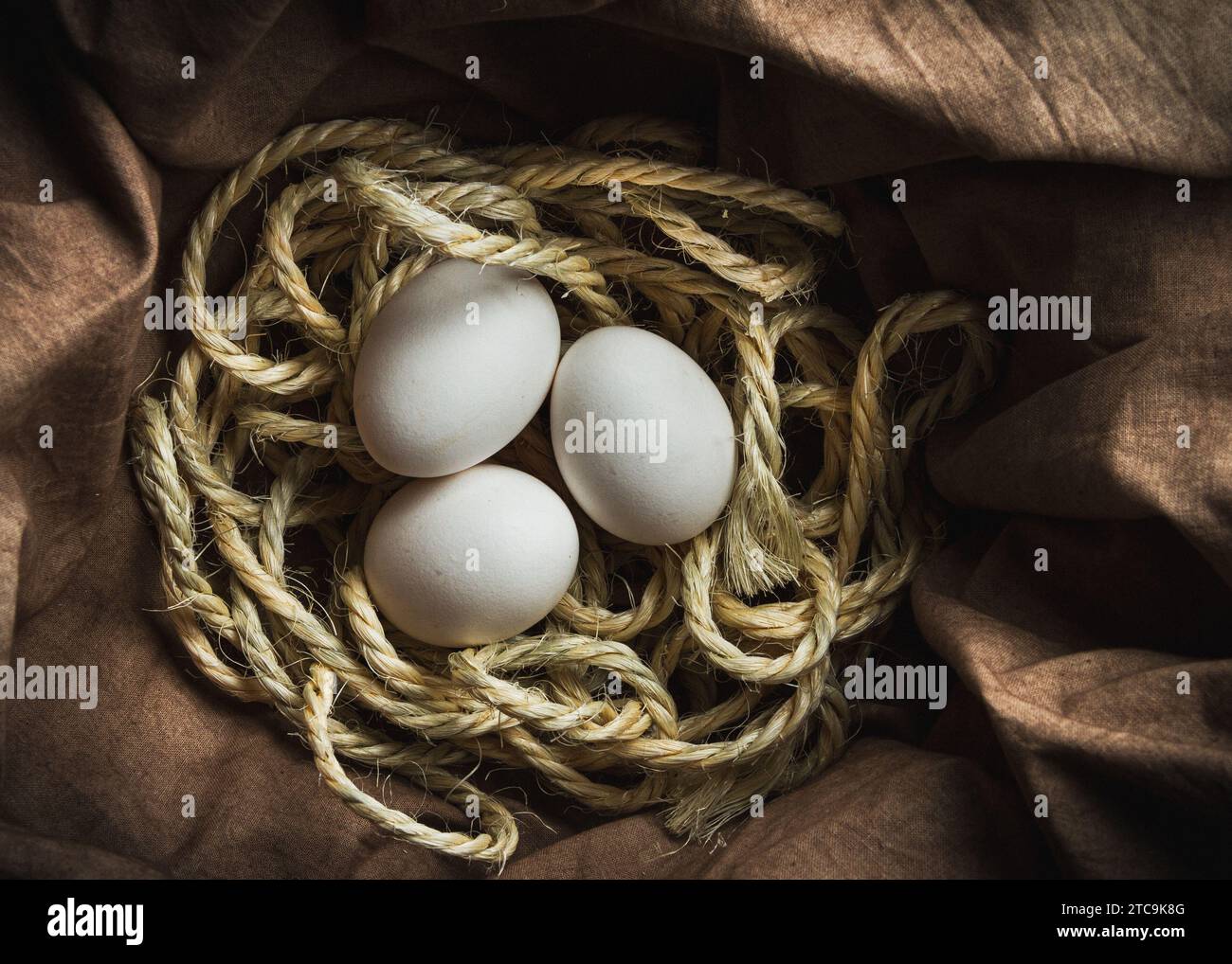 tre uova bianche riposate in un nido di corda sedute su un pezzo di stoffa marrone Foto Stock