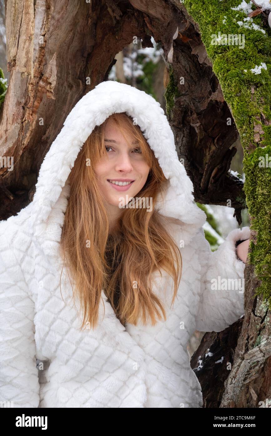 Ritratto di una bella sexy seducente giovane donna sorridente felice fuori vacanza nella foresta di alberi naturali nel freddo inverno innevato con cappotto bianco con cappuccio Foto Stock