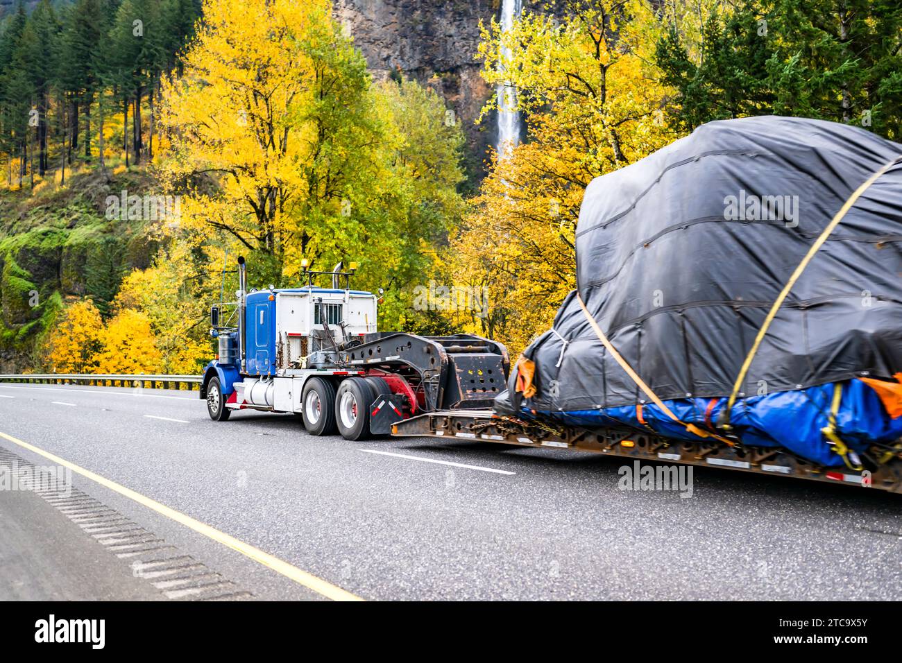 Grande carro di perforazione blu industriale per impieghi gravosi classico semicarro americano che trasporta carichi di grandi dimensioni coperti su strade autunnali con alberi gialli e W. Foto Stock