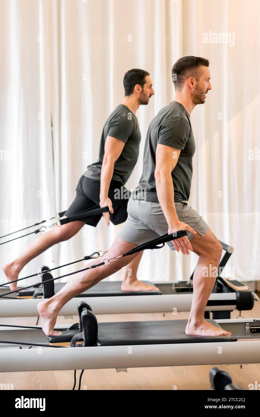 Vista laterale completa del corpo di uomini forti in abbigliamento sportivo che fanno esercizi di pilates con fasce di resistenza sui letti riformatori durante l'allenamento in palestra Foto Stock