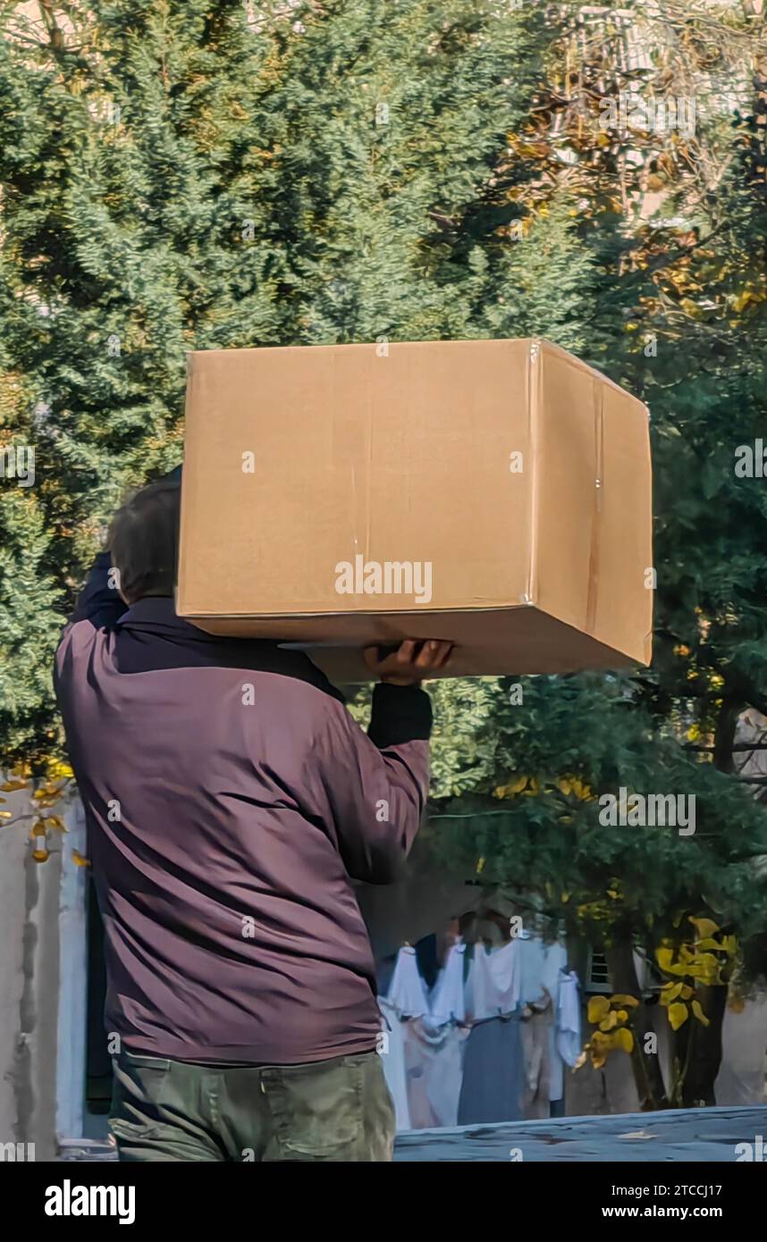 L'uomo sconosciuto con giacca marrone e pantaloni verdi porta con sé un cartoncino marrone. contro gli alberi come sfondo. Foto Stock