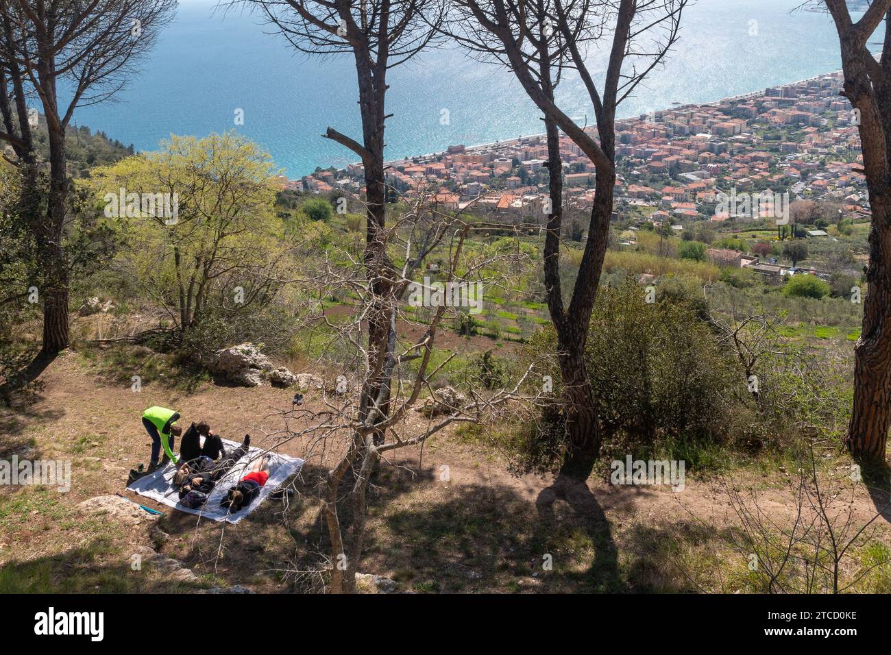 Vista sopraelevata della città costiera di pietra Ligure dalla collina di Borgio Verezzi con gli escursionisti che riposano sotto gli alberi in primavera, Savona, Liguria, Italia Foto Stock