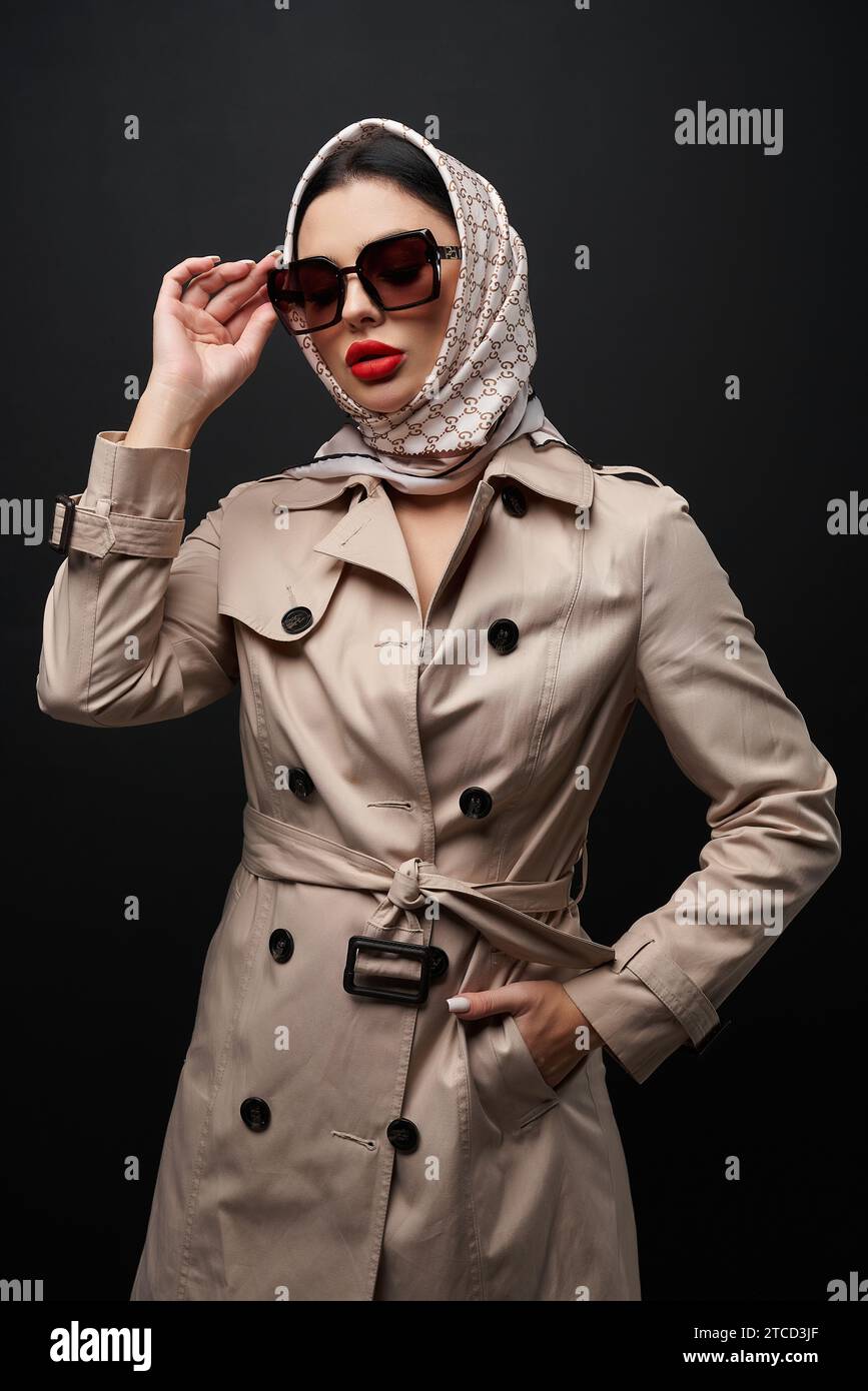 Elegante modello femminile con trench e foulard in posa per la fotocamera. Ritratto di una donna alla moda che indossa un capo, indossa occhiali da sole, mentre è in piedi Foto Stock