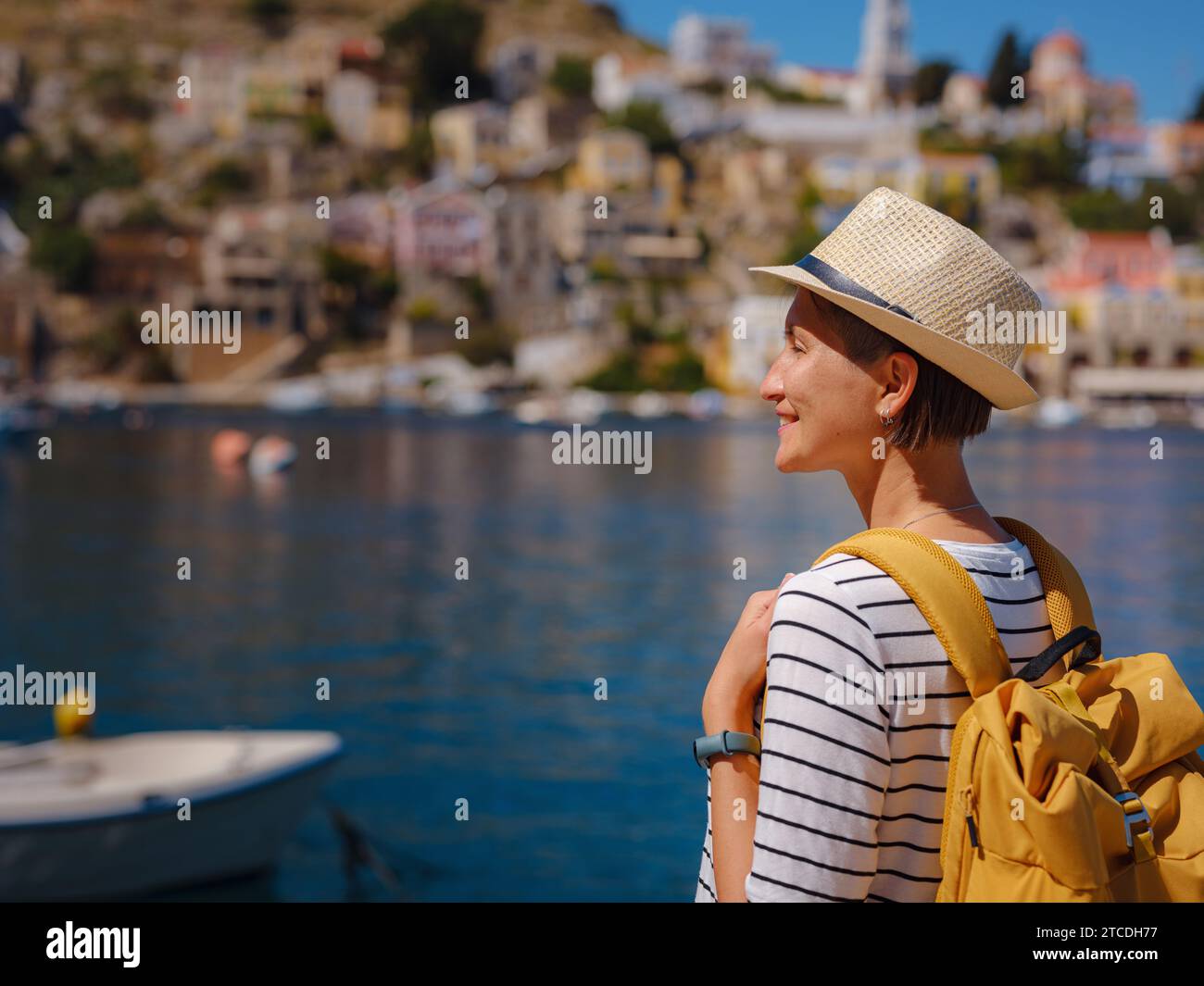 Bella asiatica felice femmina con zaino che si gode le sue vacanze alle isole Symi. Vista del porto di Symi o Simi, è una piccola isola del Dodecaneso, Grecia, atmosfera tranquilla e architettura favolosa. Foto Stock