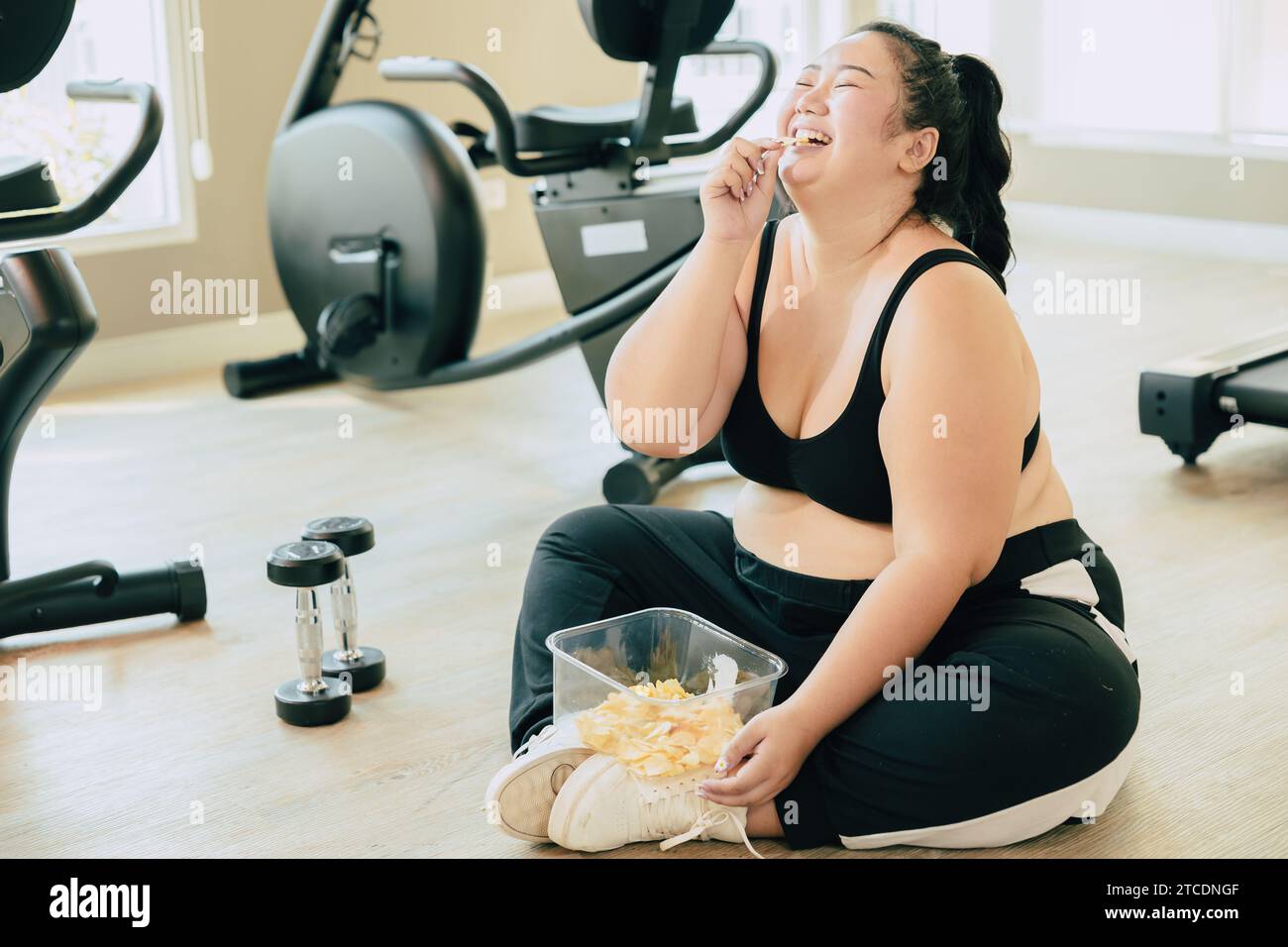 Disturbo del mangiare binge (LETTO), le donne grasse felici ridono goditi il cibo inarrestabile di patatine fritte durante il relax frenate dall'esercizio nel fitness club sportivo. Foto Stock