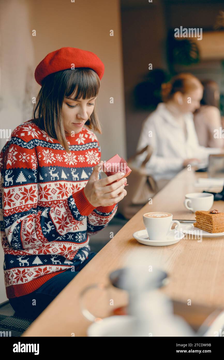 Una donna premurosa in un maglione per le vacanze scarta delicatamente un regalo, immersa nello spirito festivo di un caffè caldo e invitante Foto Stock