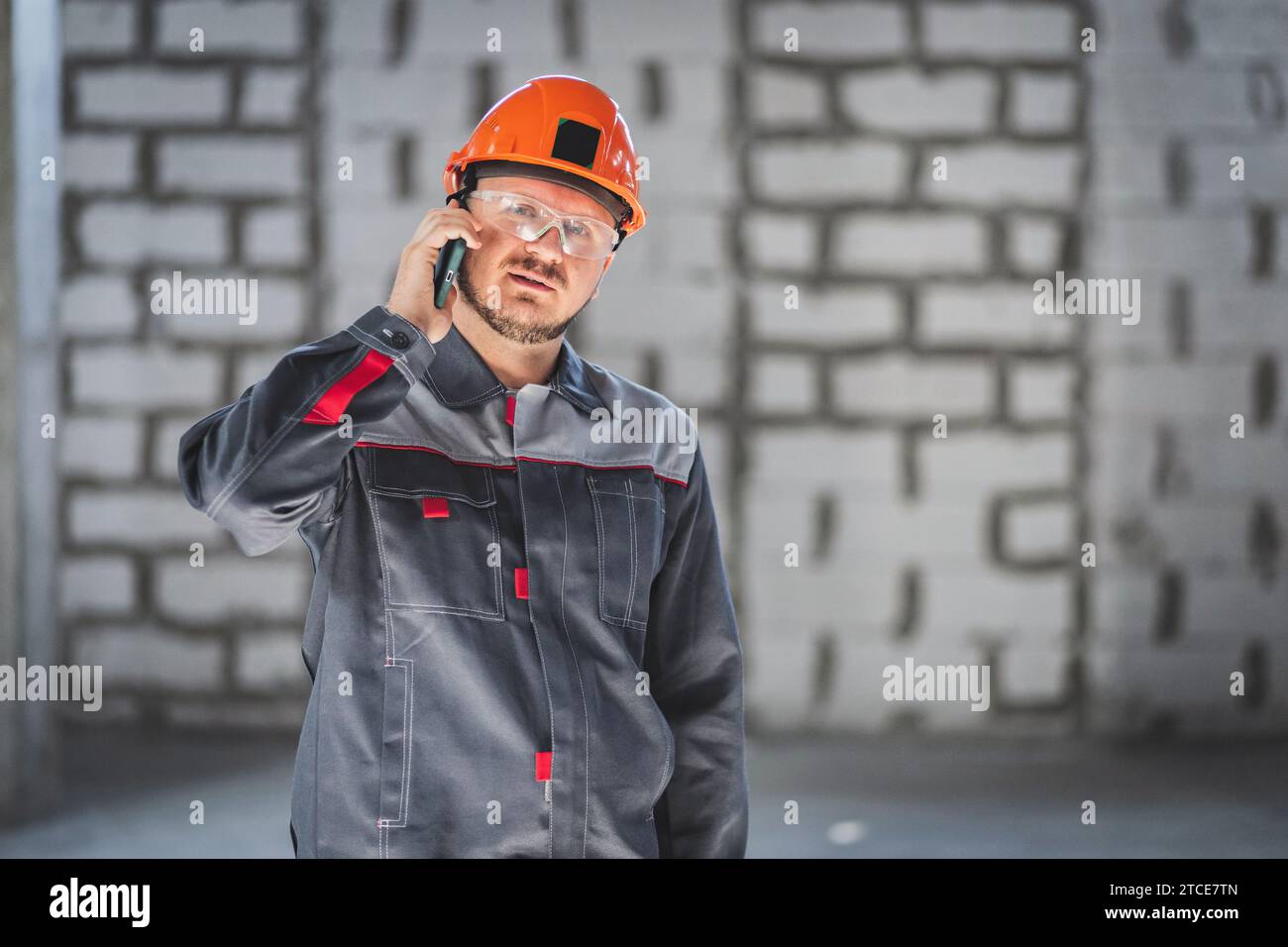 Costruzione barbuto lavoratore caucasico in tuta, casco e colle protettive che parla su smartphone in un cantiere edile Foto Stock