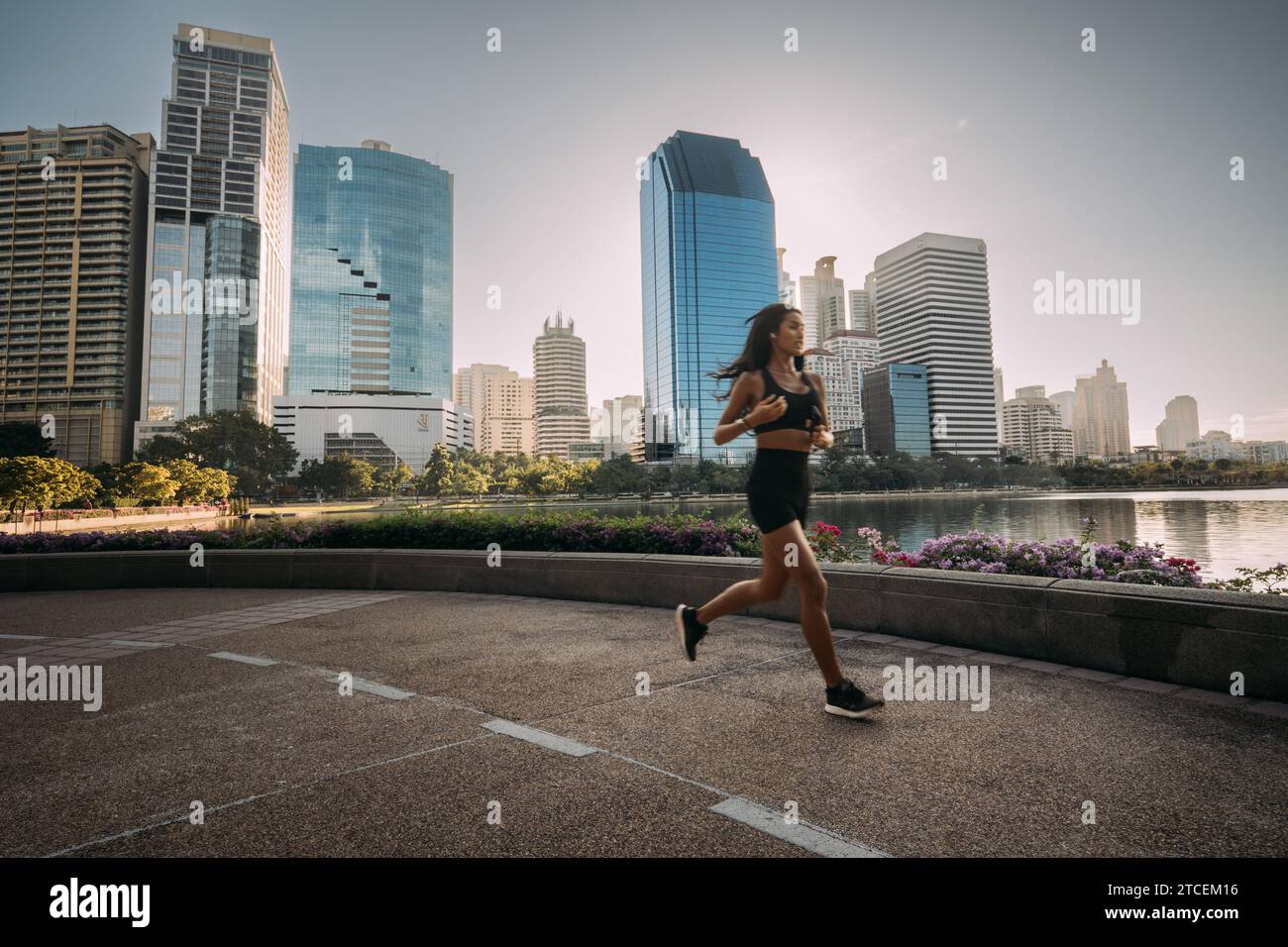 La giovane donna asiatica in costume sportivo corre su una pista vicino a un lago, con lo skyline della città sullo sfondo. Foto Stock