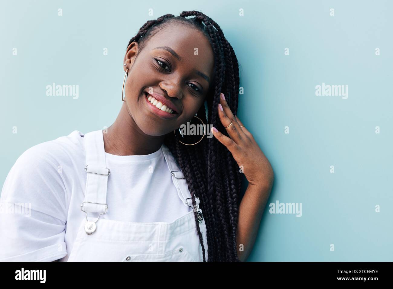 Ritratto ravvicinato di una giovane donna con trecce. Donna sorridente in abiti bianchi. Donna positiva che si appoggia a una parete blu e si regola le trecce mentre guarda Foto Stock
