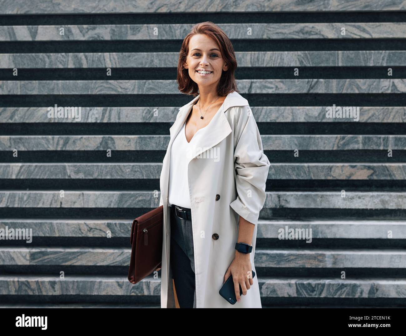 Allegra donna di mezza età con abiti eleganti che guarda la fotocamera mentre si trova all'aperto. Donna in cappotto con cartella in pelle. Donna con Ginger hai Foto Stock