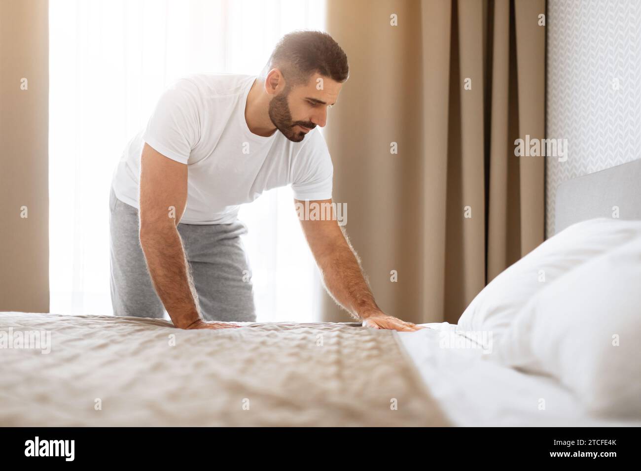 L'uomo riposato fa il suo letto dopo il sonno riordinando la camera da letto moderna Foto Stock