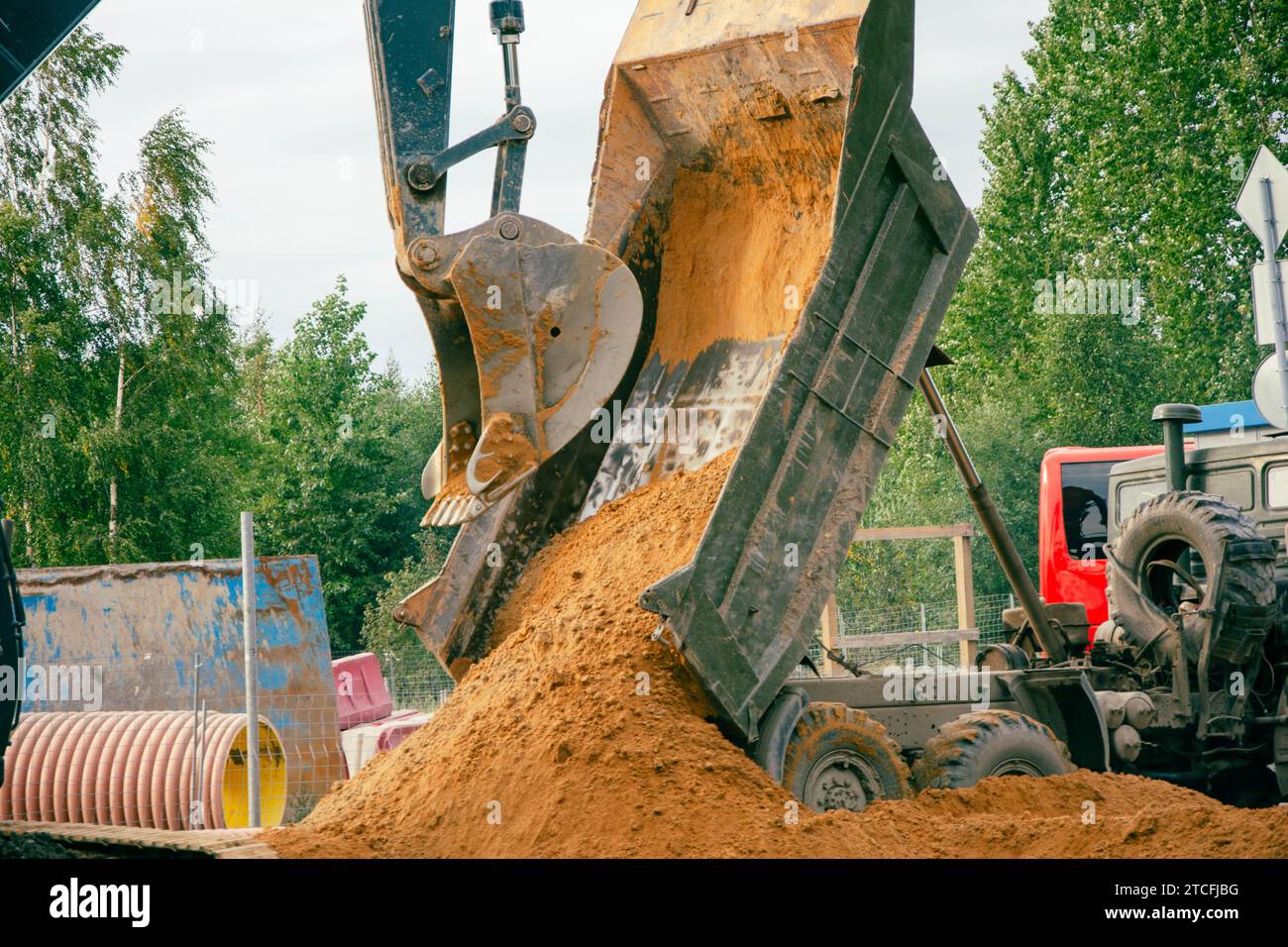 Un escavatore giallo con una grande benna sta scaricando la sabbia da un camion in un vivace cantiere in una giornata di sole, mostrando le attrezzature pesanti Foto Stock