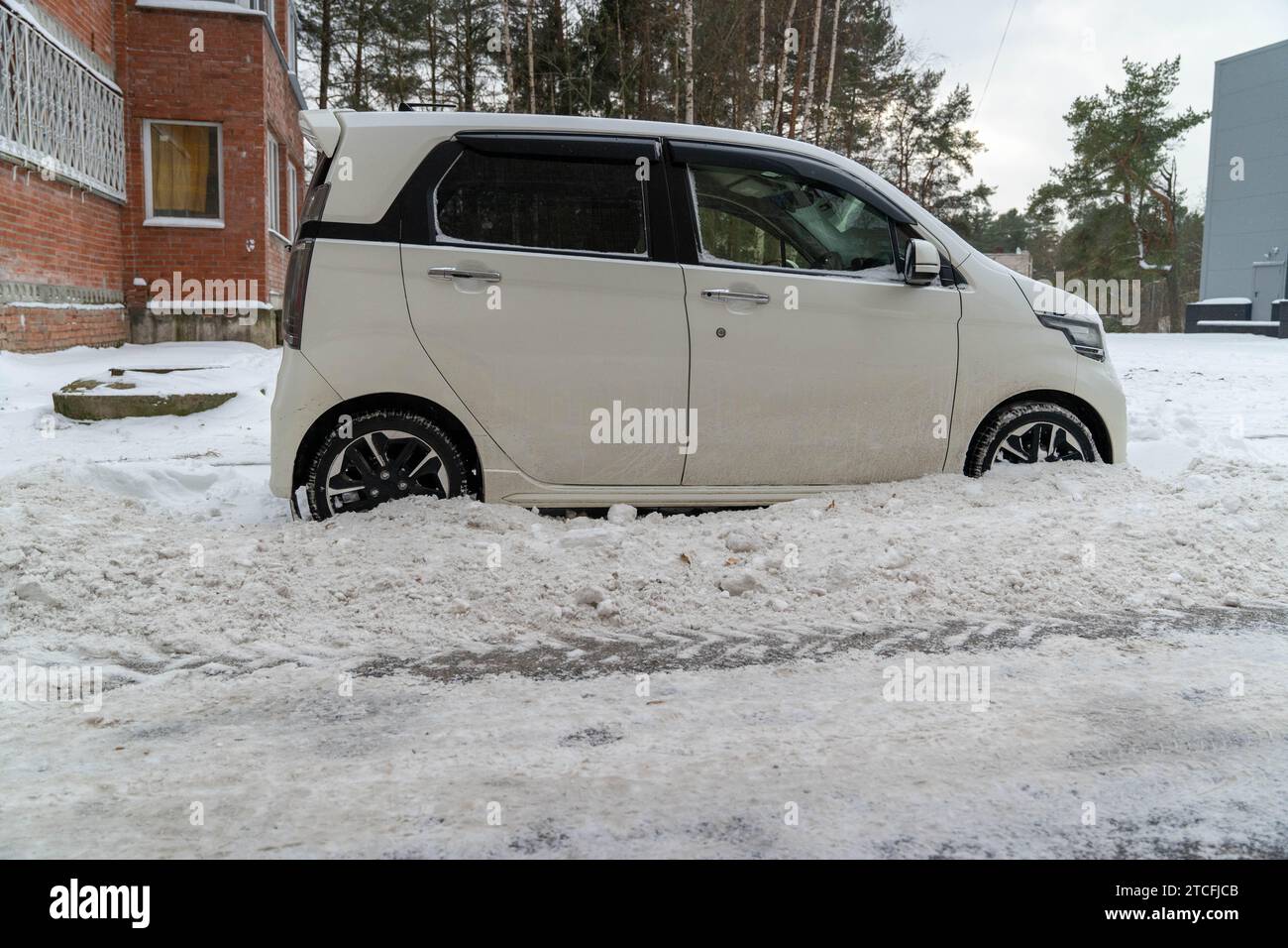 Una piccola auto coperta di detriti di neve sporca viene parcheggiata su una strada cittadina dopo la rimozione della neve. Il veicolo è circondato da grandi cumuli di neve, creando una W. Foto Stock