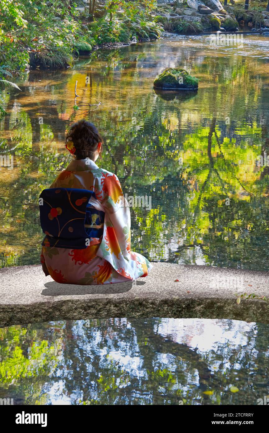 Una giovane donna giapponese in kimono in un giardino giapponese seduta a contemplare gli specchi sull'acqua dello stagno Foto Stock