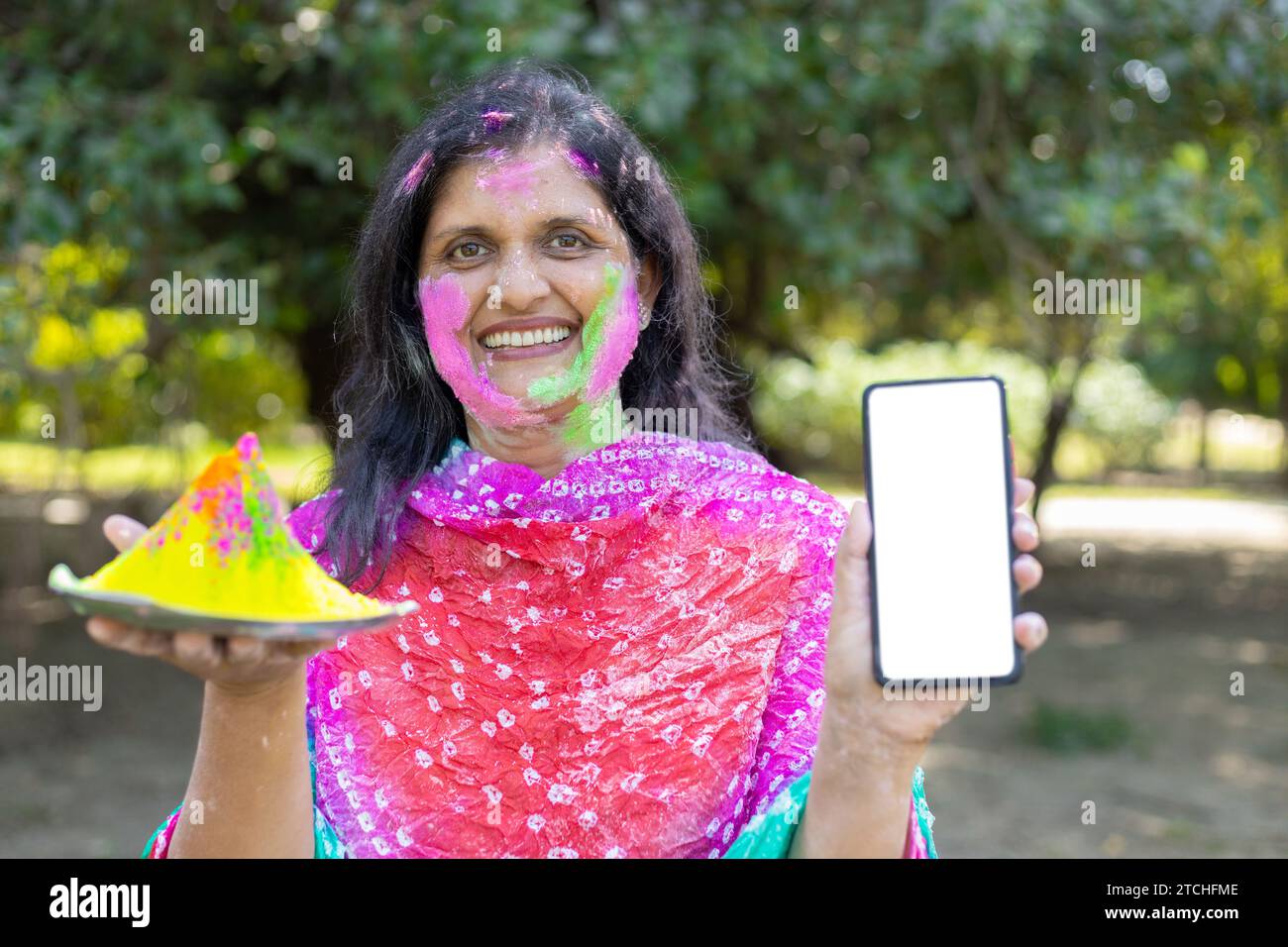 Donna indiana felice che indossa kurta bianca con smartphone e piatto di polvere colorata o gulal che celebra il festival holi al parco o in giardino. Mockup, poliziotto Foto Stock