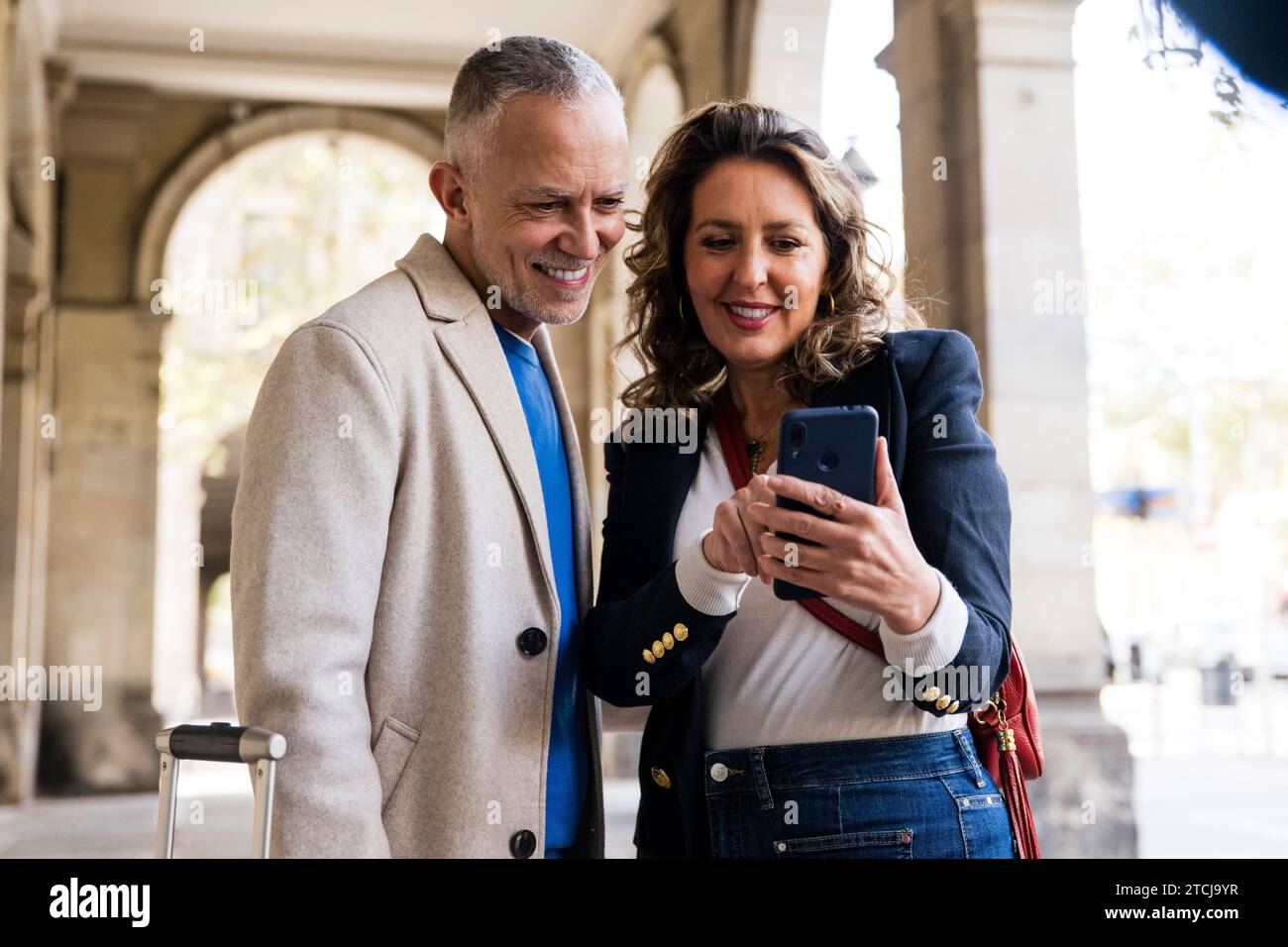 Coppia di adulti di medie dimensioni che usa lo smartphone insieme per strada. Marito e moglie anziani sorridenti che scattano una foto in una città. Foto Stock