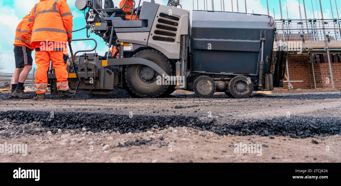 Asfaltatrice riempita con asfalto caldo che posa una nuova superficie stradale sul nuovo sito di sviluppo di abitazioni residenziali e operatore stradale in arancione hi-viz Next Foto Stock