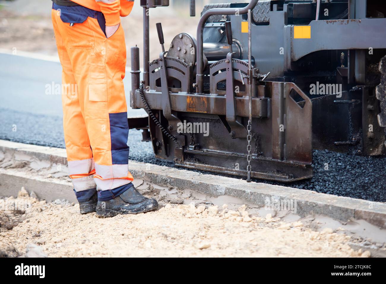 Asfaltatrice riempita con asfalto caldo che posa una nuova superficie stradale sul nuovo sito di sviluppo di abitazioni residenziali e operatore stradale in arancione hi-viz Next Foto Stock