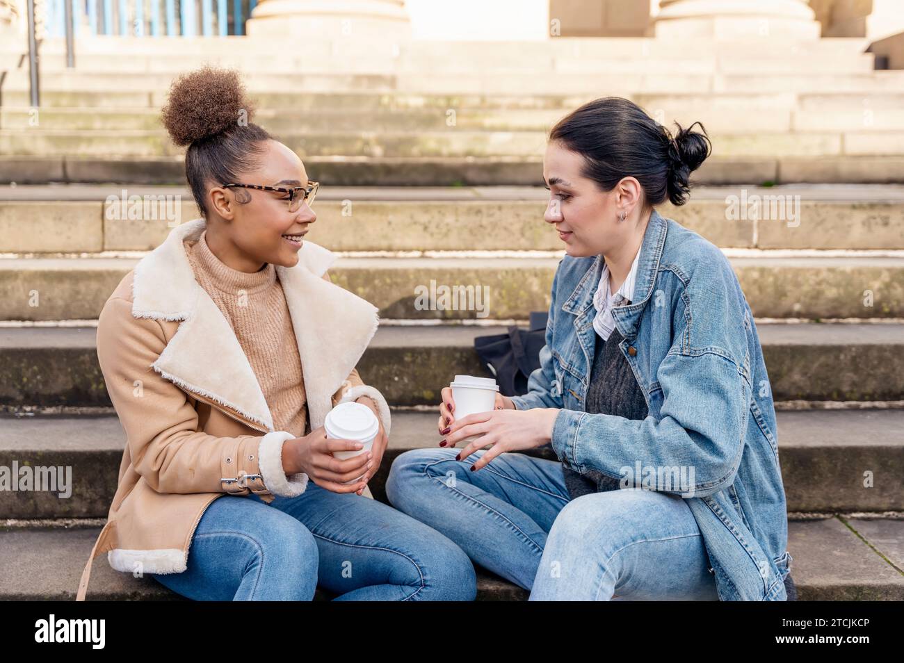 due studentesse in una giacca di denim parlano tra loro, al telefono, ridono, bevono caffè sulle scale in città. Foto Stock