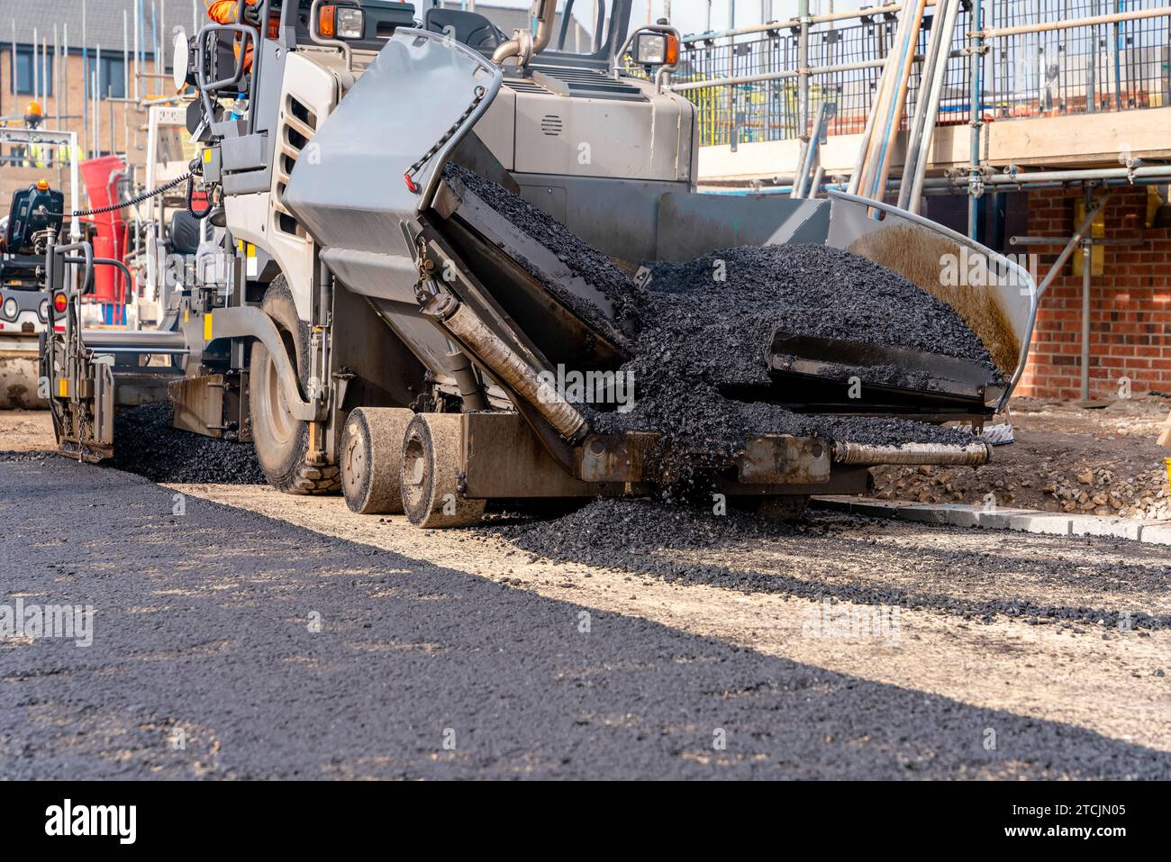 Asfaltatrice riempita con asfalto caldo che posa una nuova superficie stradale sul nuovo sito di sviluppo di abitazioni residenziali e operatore stradale in arancione hi-viz Next Foto Stock