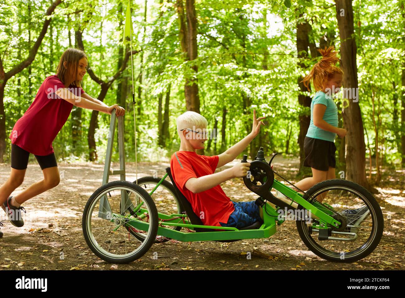 Ragazza giocosa che spinge un ragazzo disabile in bicicletta reclinata sul sentiero della foresta Foto Stock