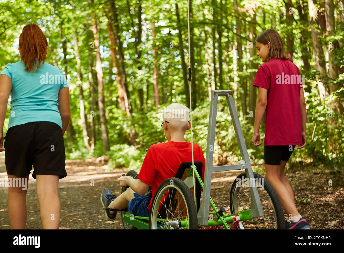 Vista posteriore delle ragazze che camminano con un bambino portatore di handicap in bicicletta reclinata nella foresta Foto Stock