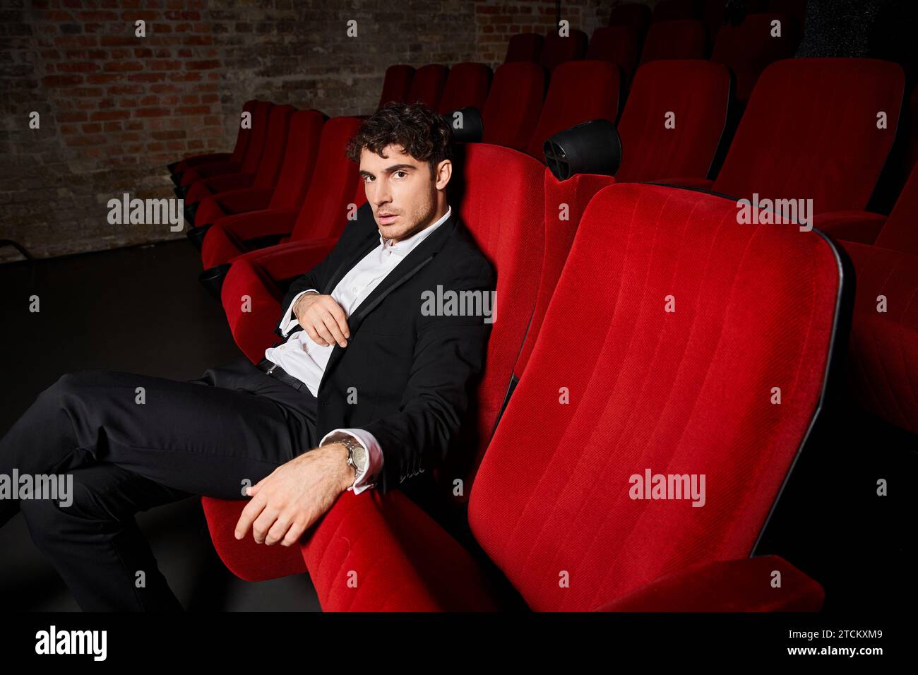 affascinante uomo in elegante vestito nero seduto su sedie rosse al cinema e guardando la macchina fotografica Foto Stock