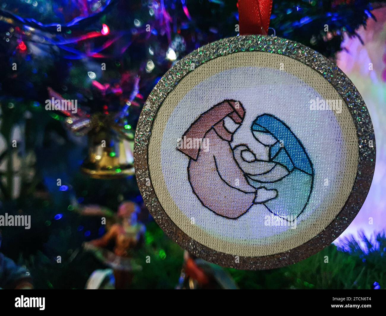 La Sacra famiglia dipinta e ricamata con un ciondolo che spicca sull'albero di Natale con luci colorate. Foto Stock