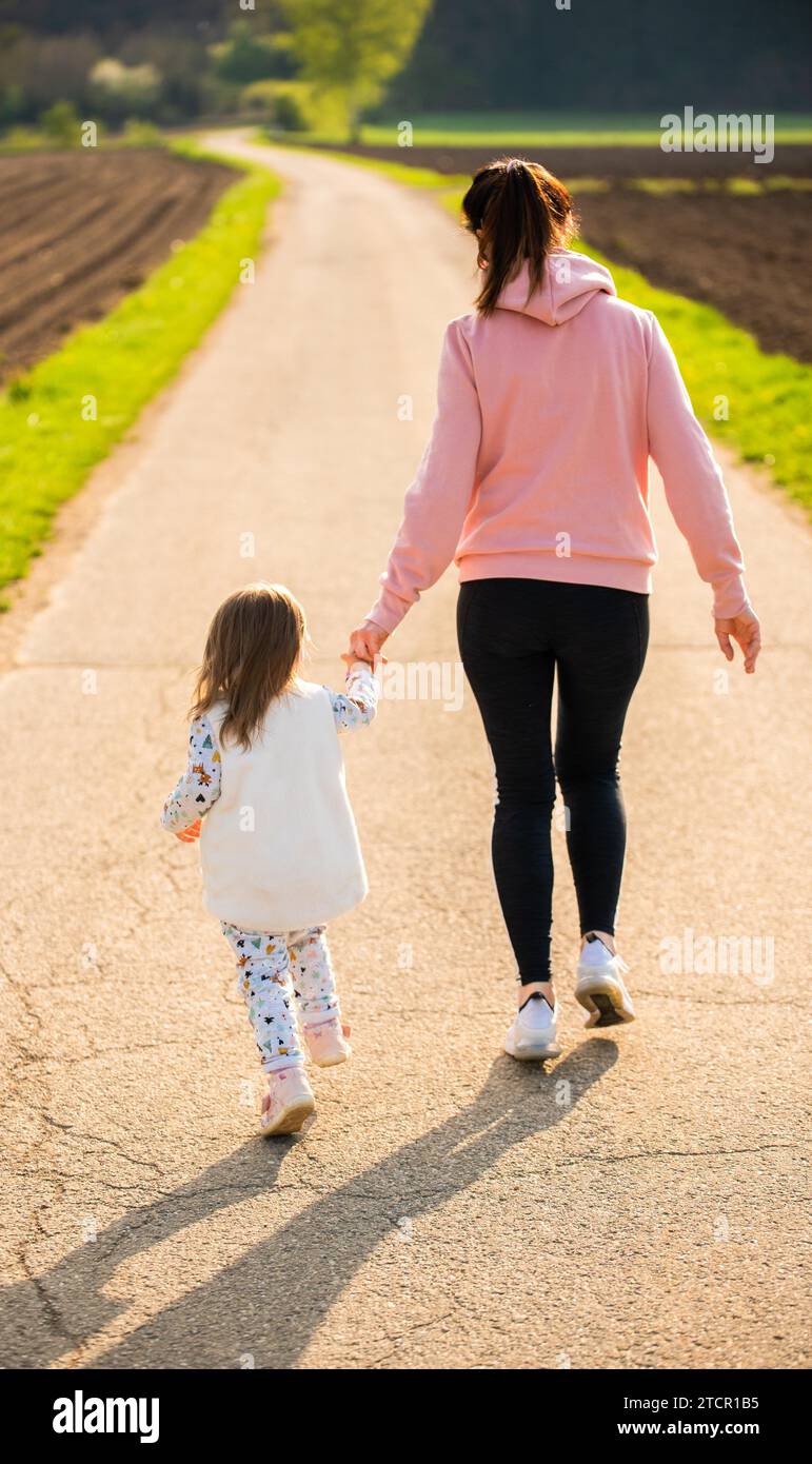Madre e bambino camminano su una strada di campagna tra campi agricoli verso la foresta nel vilage austriaco del tramonto. Vita familiare attiva senza Foto Stock