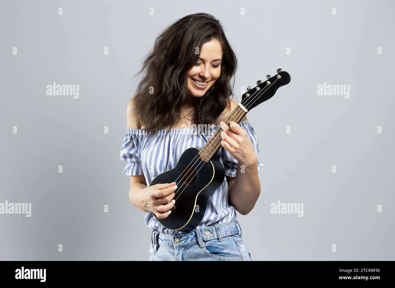 Attraente donna bruna che gioca a ukulele, isolata su sfondo grigio Foto Stock