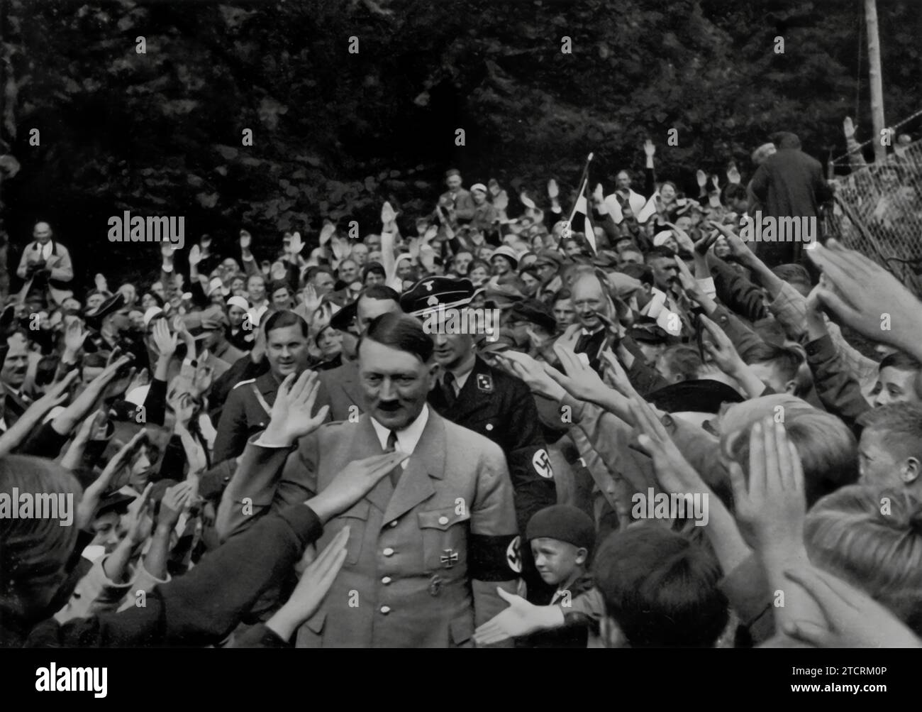 Adolf Hitler viene catturato camminando attraverso una folla fuori dalla sua casa di campagna a Obersalzberg. Questa immagine lo ritrae mescolandosi con le persone in un modo apparentemente avvicinabile, in contrasto con il suo personaggio pubblico spesso autorevole. La presenza di una folla nel suo ritiro privato, il Berghof, significa la fusione di aspetti personali e politici della sua vita. La fotografia potrebbe essere destinata a mostrare Hitler come un leader popolare, accessibile e collegato al popolo, rafforzando la sua immagine di figura carismatica. Foto Stock