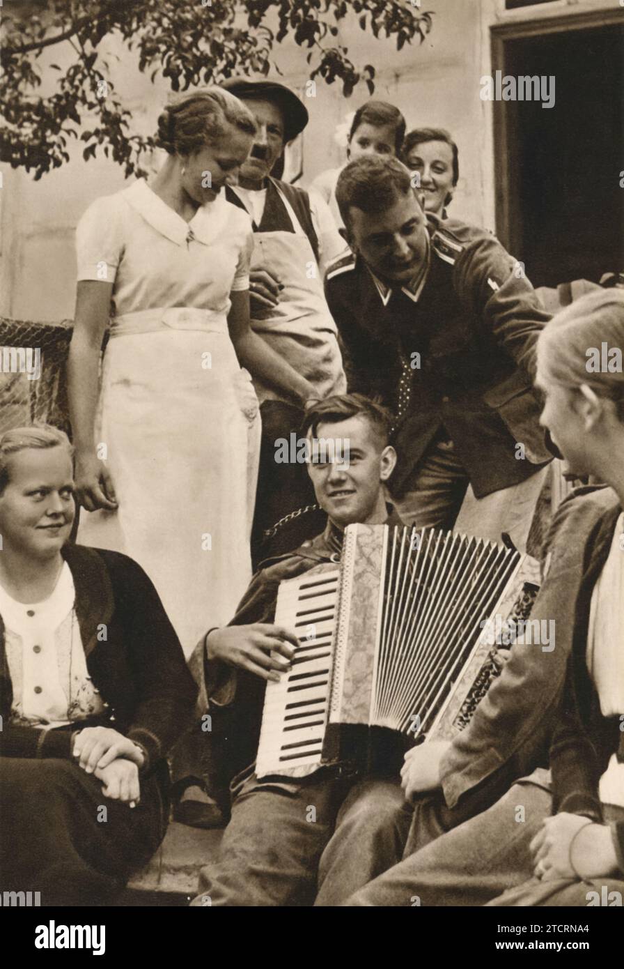 Nei quartieri, un soldato tedesco suona una fisarmonica, creando un'atmosfera vivace e piacevole. Questa scena cattura un momento di svago e di cameratismo tra le truppe, un netto contrasto con i rigori della vita militare. La musica e il divertimento condiviso sottolineano l'importanza del morale e dei momenti più leggeri che aiutano a sostenere i soldati in tempi difficili. Foto Stock
