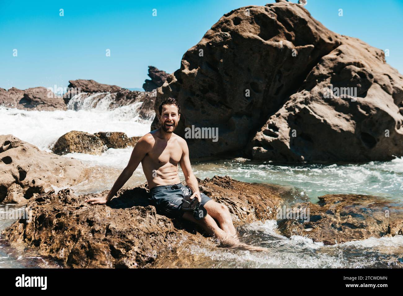 Uomo seduto sulle rocce nell'acqua alla spiaggia del Monte Maunganui, nuova Zelanda. Foto Stock