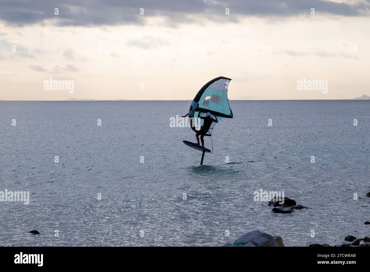 Un'imbarcazione da surf in mare calmo a tutta velocità. Con una vela verde e alcune rocce in acqua in primo piano Foto Stock